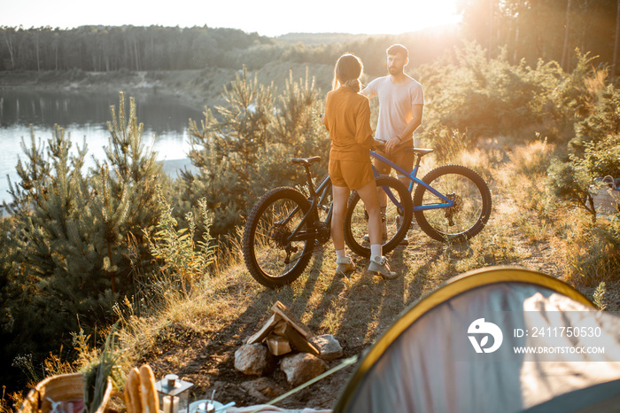 Young couple standing with mountain bicycles at the campsite, traveling in the forest near the lake on the sunset