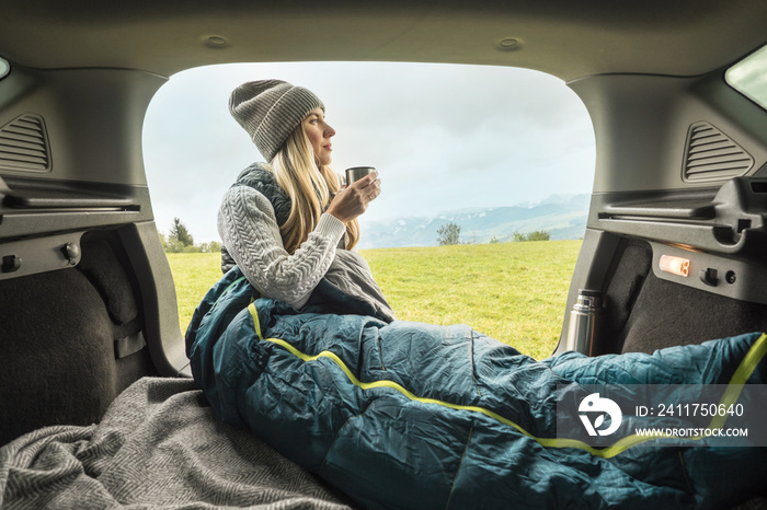 Girl resting in her car. Woman hiker, hiking backpacker traveler camper in sleeping bag, drinking hot tea and relaxing on top of mountain. Health care, authenticity, sense of balance and calmness.