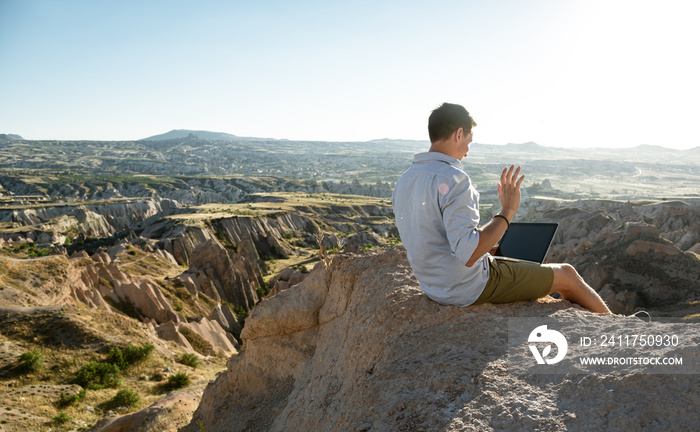 Young man sitting at the edge of valley and making video call with laptop. Copy space