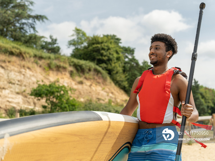 Young man carrying paddleboard and oar on beach