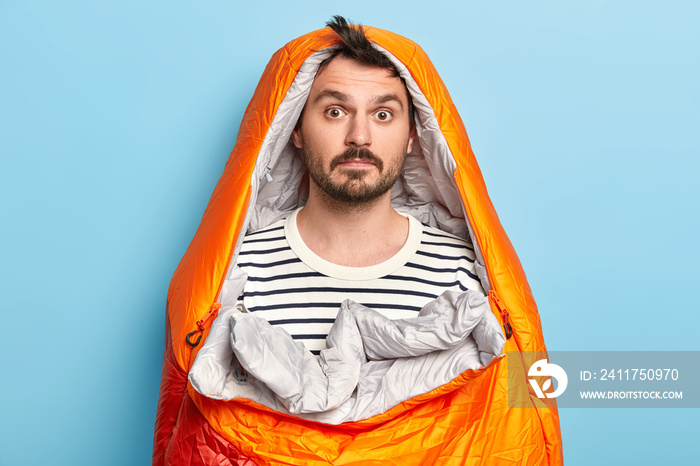 Photo of surprised bearded male camper poses in sleeping bag, wears striped jumper, spends weekend actively, poses over blue background, has expedition near rocky mountains. Active rest concept