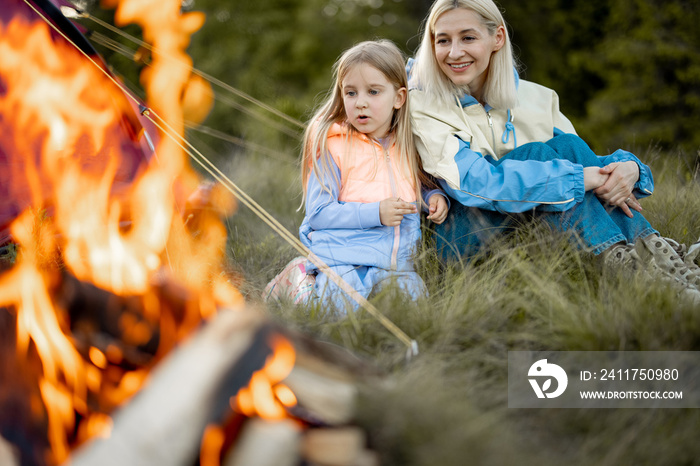 Young woman with her little girl sit by the campfire, bonding together while travel on nature. Mother with daughter warming up at campsite