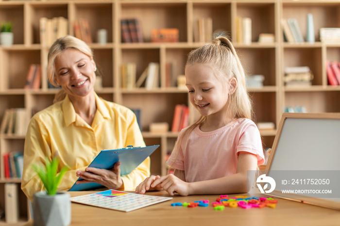 Cute little caucasian girl exercising at private preschool class, sitting at table with teacher and doing various tests