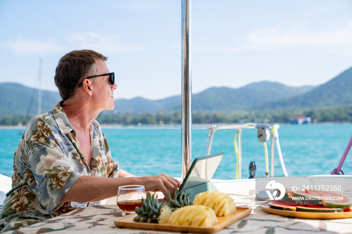 Caucasian businessman working on digital tablet for online corporate business while catamaran boat sailing on summer vacation. Handsome man enjoy outdoor lifestyle activity tropical travel trip