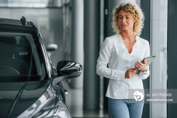 Tablet is in the hands. Woman with curly blonde hair is in autosalon