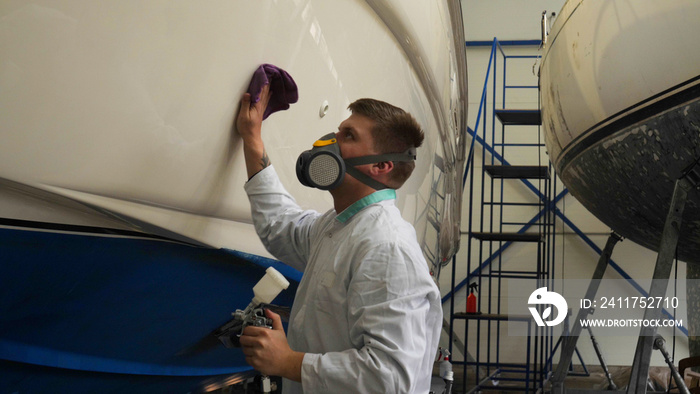 A young man (guy) is a specialist in cleaning the yacht, in a respirator and using a spray on the background of the yachts. Concept from: Professional, Specialist, Cleaning, Yacht service, Preparation