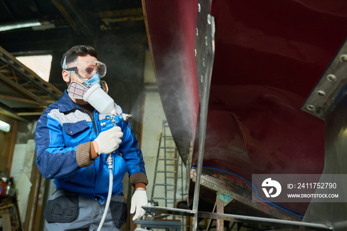 Low angle portrait of unrecognizable worker wearing protective mask painting boat using paint sprayer in yacht workshop, copy space