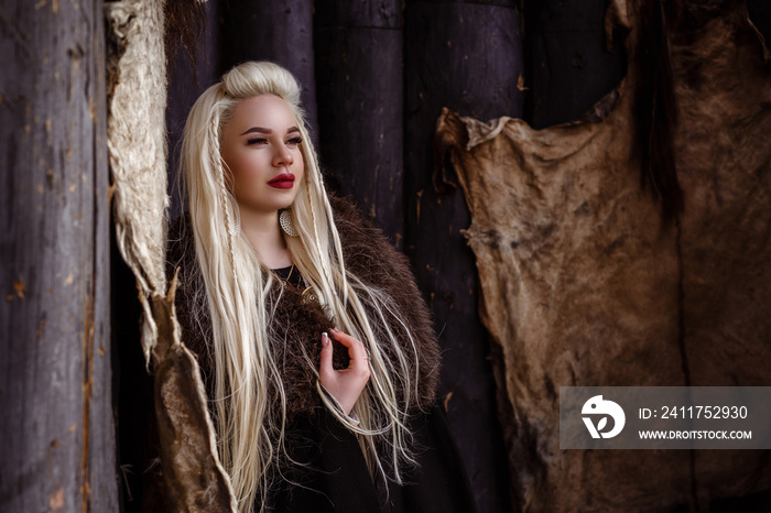 Outdoors portrait of beautiful furious scandinavian warrior ginger woman in a traditional clothes with fur collar, with sword in her hand and wooden Viking Village view on the background.