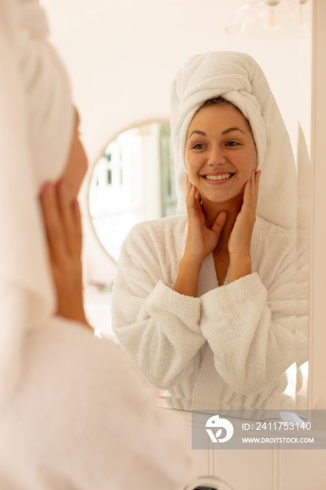 Happy caucasian woman wearing robe, looking at her reflection in mirror and smiling in bathroom