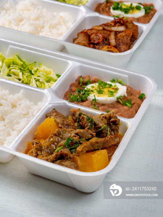 food delivery, styrofoam lunch box with beef meat cooked with cassava, zucchini, rice, beans and boiled egg on white wood texture.