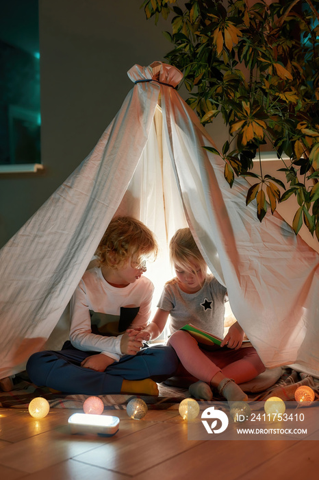 Cozy house. Two little siblings, boy and girl reading a book together while sitting on a blanket in a hut made with bedsheets at home