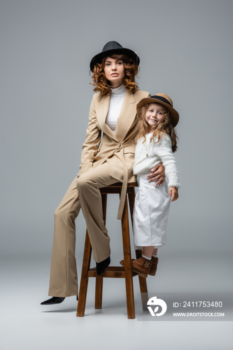 elegant mother and daughter in white and beige outfits posing on chair on grey