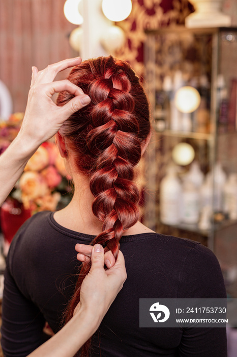 Beautiful, red-haired girl with long hair, hairdresser weaves a French braid, in a beauty salon. Professional hair care and creating hairstyles.