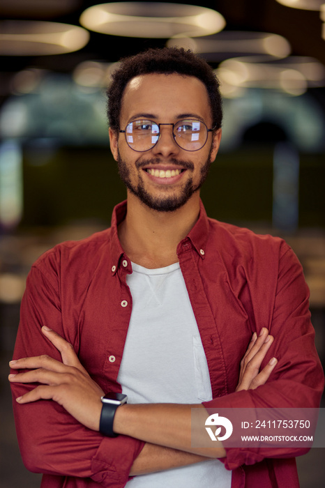 Vertical shot of a young happy mixed race businessman or office worker in casual wear keeping arms crossed, looking at camera and smiling while posing in the office