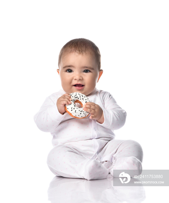 Happy laughing infant baby toddler in white onepiece overall sits on the floor holding donut in hands going to bite