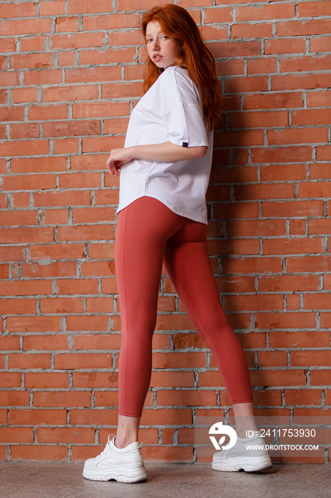 Fashion style studio portrait of beautiful red-haired young girl in leggings pants and white t-shirt. Model standing and posing beside brick wall