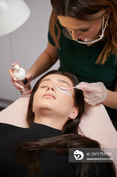 Close up of beauty specialist in sterile gloves applying cleansing mousse on woman eyelashes with cosmetic lash brush. Eyelash stylist preparing woman lashes for applying extensions.