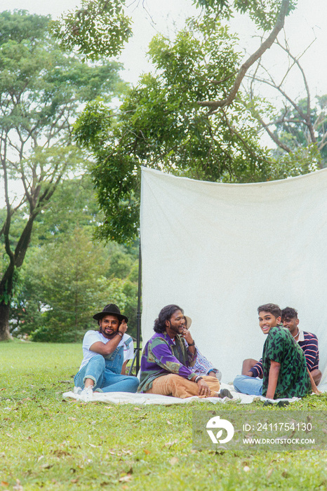 Malaysian Indian men in a group against a cloth backdrop in a park surrounded by trees, talking, laughing and sitting together