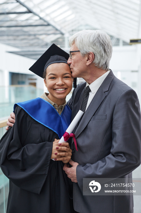 Vertical portrait of proud father kissing daughter during graduation ceremony in college