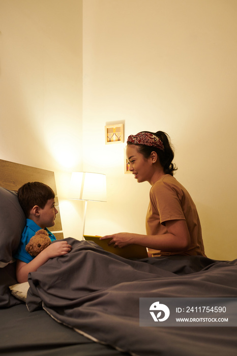 Smiling mother reading book for her preteen son lying in bed with teddy bear