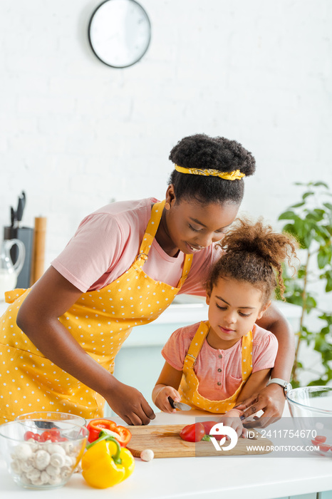 happy african american woman cutting bell pepper with cute daughter