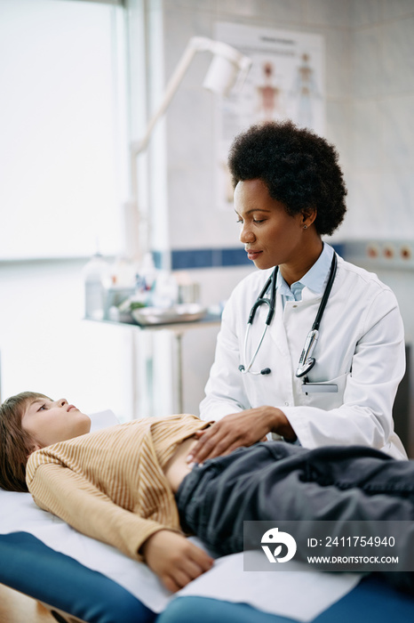 Black female pediatrician examining abdomen of small boy at doctor’s office.