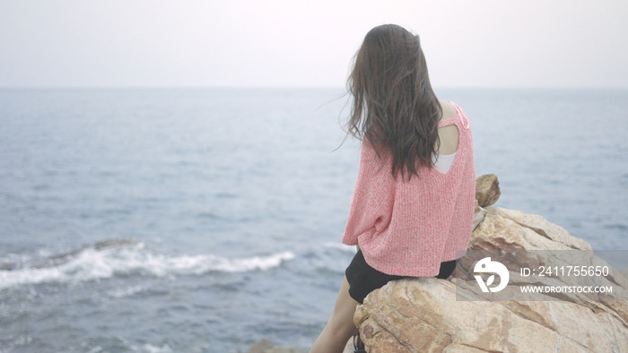 A beautiful woman in pink sweater sits on a rock above the sea, looking at the ocean. Girl traveler, backdrop of the sea and sky. thinking, freedom, solitude, relaxed, Enjoying concept. copy space.