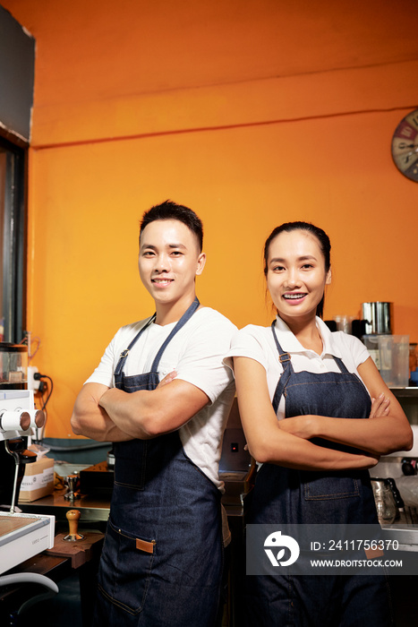 Asian young couple of bartenders standing in aprons and smiling at camera while working in coffee shop