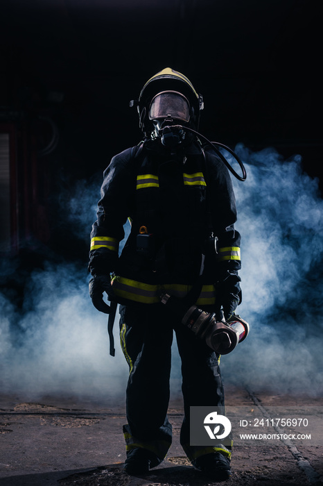 Portrait of a female firefighter wearing a helmet and all safety equipment a while holding a tomahawk and wearing an oxygen mask indoors surrounded by smoke
