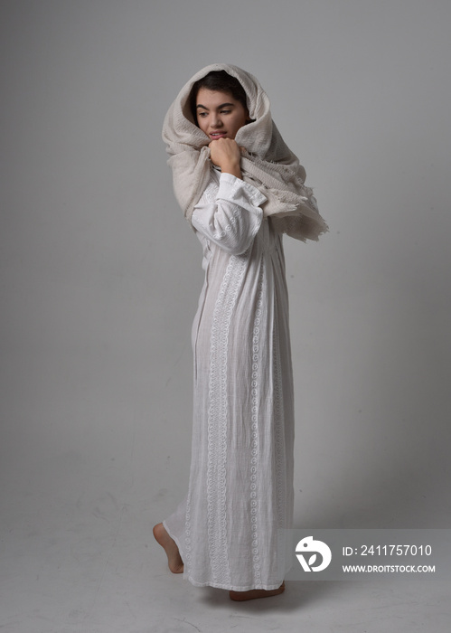 Full length portrait of young woman wearing classical white gown and a head covering veil in biblical style, sitting pose on light studio background.