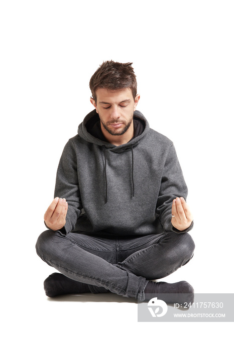 Hombre joven aislado sentado meditando con las piernas cruzadas en el piso sobre fondo blanco, con sudadera gris, jean gris y medias grises