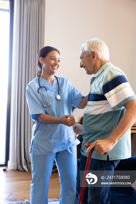 Biracial female physiotherapist assisting caucasian senior man in standing with walking cane at home