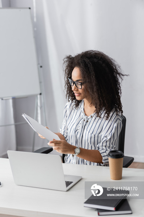 happy african american freelancer in glasses holding folder near laptop and paper cup on desk