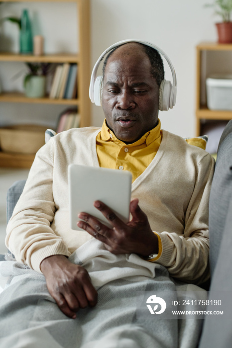 Senior African man in wireless headphones watching video on digital tablet during his rest on sofa at home