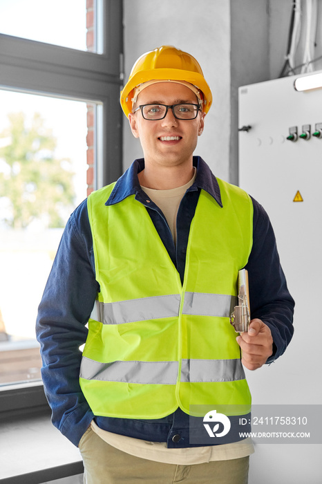 construction business and building concept - happy smiling male electrician or worker in helmet and safety west with papers on clipboard and pencil at electric board