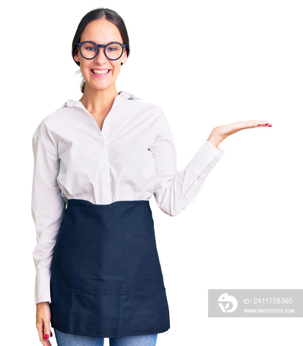 Beautiful brunette young woman wearing professional waitress apron smiling cheerful presenting and pointing with palm of hand looking at the camera.