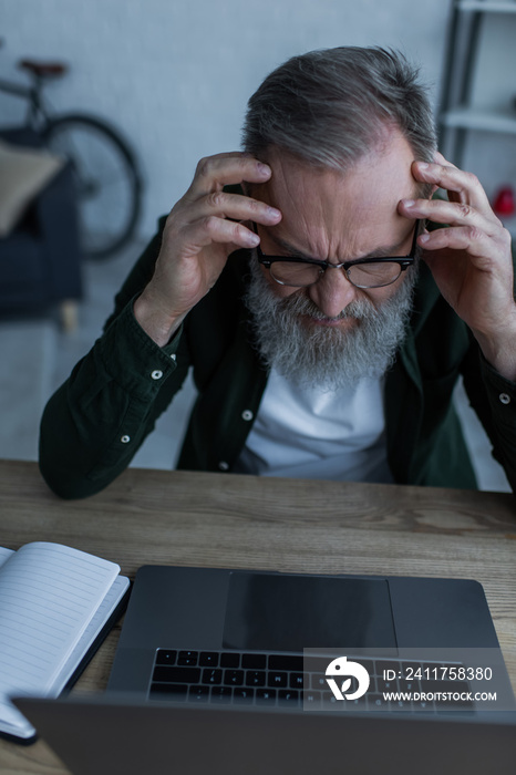 stressed senior man in eyeglasses frowning near laptop at home.