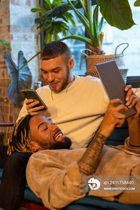 Male couple using smart phone and tablet on sofa