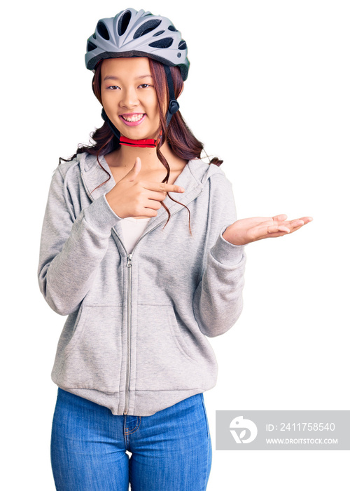 Young beautiful chinese girl wearing bike helmet amazed and smiling to the camera while presenting with hand and pointing with finger.