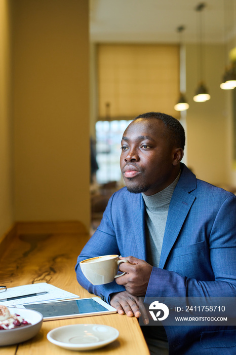 African serious businessman sitting at table with tablet pc and looking pensive while drinking coffee in cafe