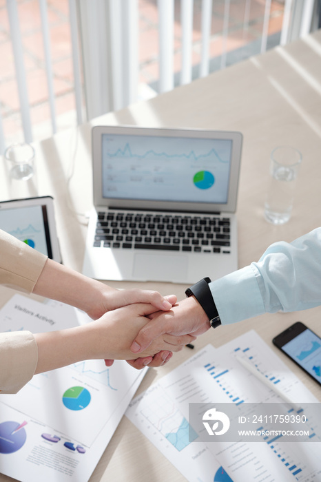 Hands of business people shaking hands over table with various charts and reports after having successful meeting