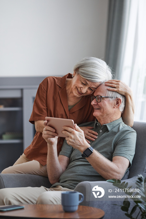 Vertical portrait of happy senior couple using digital tablet together at home and embracing