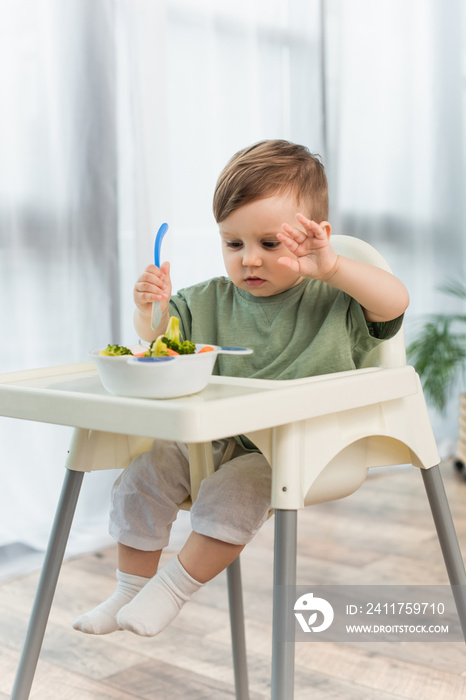 Toddler boy holding spoon near blurred bowl of vegetables on high chair