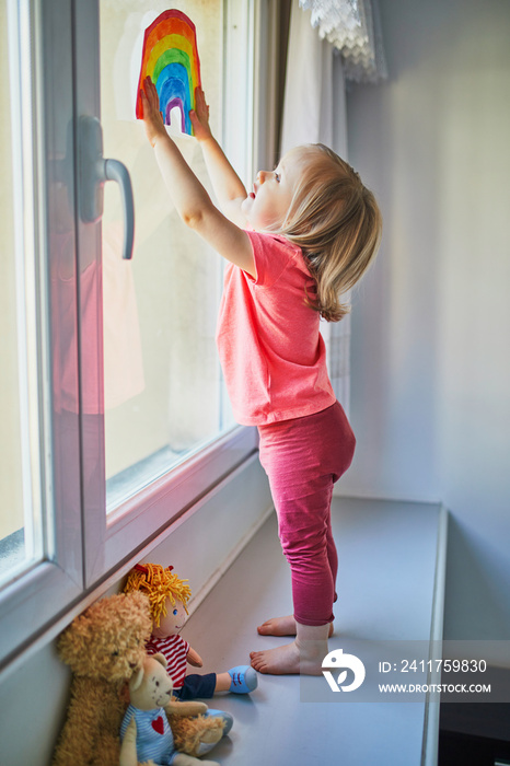 Adorable toddler girl attaching drawing of rainbow to window glass