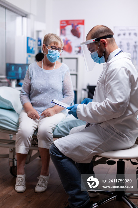 Doctor reading diagnosis of senior woman illness wearing face mask against covid pandemic as safety precaution in hospital. Healthcare medical physician consultation during COVID-19 global crisis.