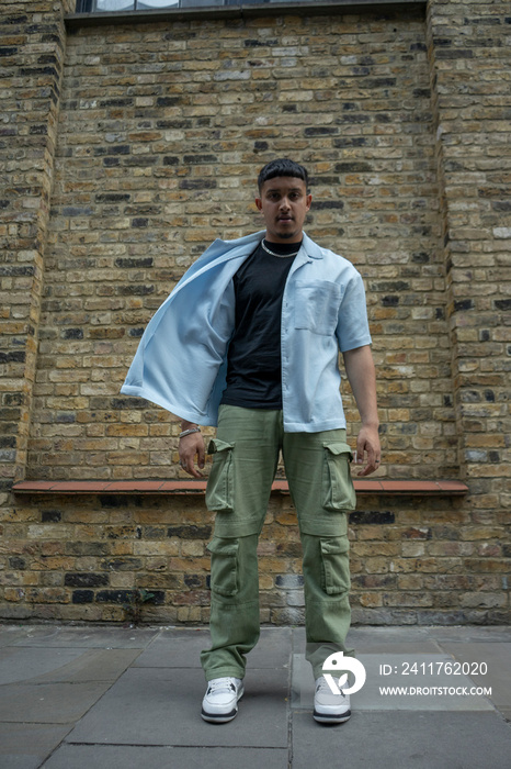 Man wearing unbuttoned shirt and cargo pants standing in front of brick wall