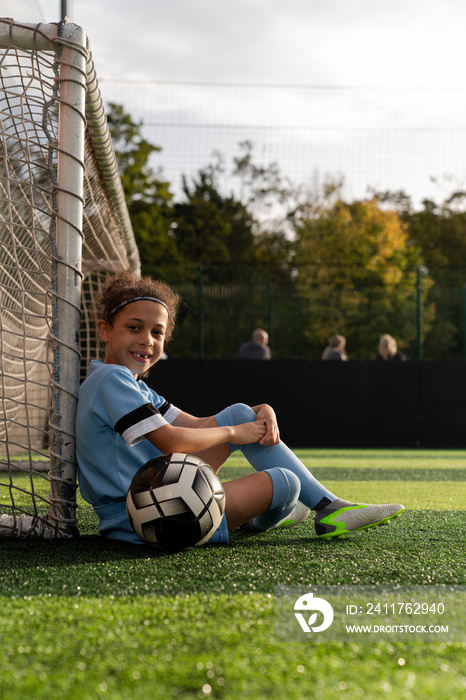 Portrait of girl (6-7) with ball sitting next to soccer goal