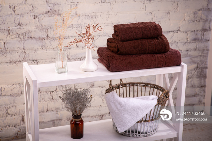 Rack with a stack of three brown color towels and baskets with clean white towels and toilet decor near a brick wall. Shelf with terry towels and decorative vases. Shelving unit with clean towels