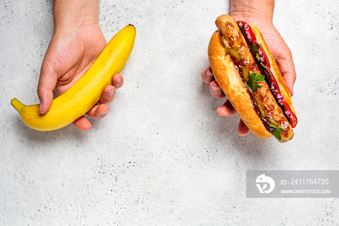 Choosing healthy food and unhealthy food concept. Vegan vs. Meat food. Banana and hot dog in hands.