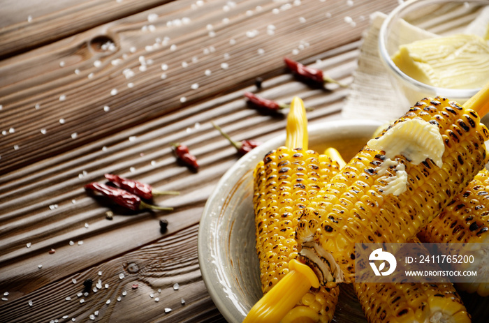 Wooden table with deep grilled sweet corn cobs under melting butter with plastic holder on clay dish